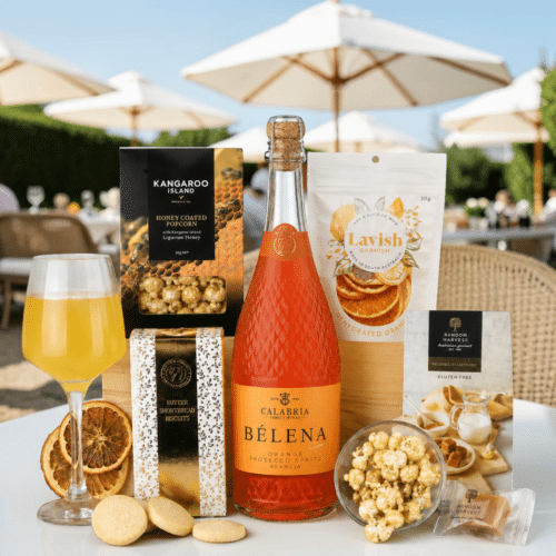 Sparkling orange Prosecco bottle surrounded by snacks (popcorn, biscuits, chocolate) and a Lavish Garnish packet on a sunny outdoor table with umbrellas in the background.