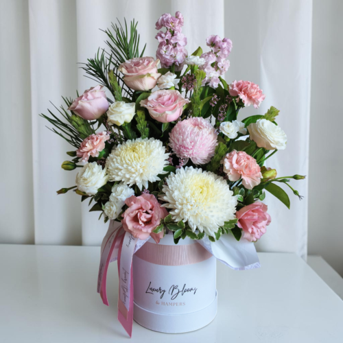 Pastel bouquet of pink and white roses, carnations, and chrysanthemums in a pale pink hatbox with ribbons, against a light backdrop.